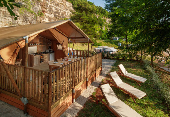 Luxe glamping-tent met houten terras, keuken en ligstoelen, gelegen in een groene, rotsachtige omgeving.