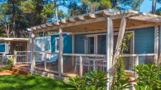 Marine Home Chalet mit Holzterrasse, weißen Geländern und Gartenmöbeln, umgeben von Pinien und grünem Rasen.