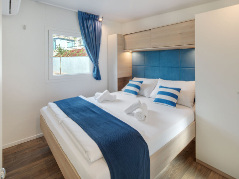 Chambre de glamping moderne avec lit double, touches bleues et blanches, sol en bois et fenêtre.