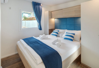 Chambre de glamping moderne avec lit double, touches bleues et blanches, sol en bois et fenêtre.