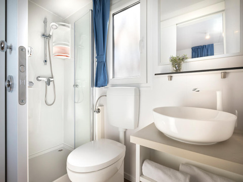 Modern glampingbadkamer met douche, toilet, wastafel, spiegel en raam met blauwe gordijn.