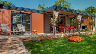 Maro Home Chalet mit orangefarbenen Wänden, Terrasse, Liegestühlen und grüner Wiese bei Sonnenschein.