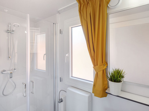 Salle de bain lumineuse avec douche, rideau jaune et plante sur la fenêtre au chalet Maro Home.