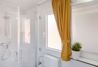 Salle de bain lumineuse avec douche, rideau jaune et plante sur la fenêtre au chalet Maro Home.