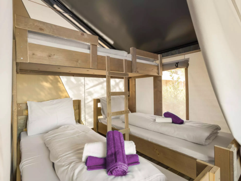 Interno di una tenda safari con letti a castello, due letti singoli, lenzuola bianche e asciugamani viola.