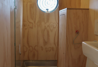 Baño compacto con paredes de madera, ventana redonda y inodoro en Wrap house de Resort de Parel, Países Bajos.