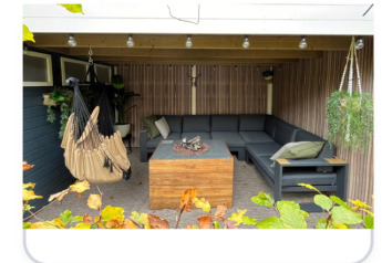 Salon de jardin couvert chaleureux avec canapé d’angle, fauteuil suspendu et brasero au Veluwe Chalet.