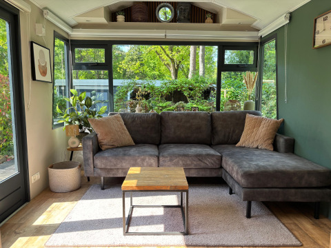 Moderne og hyggelig stue med stor grå hjørnesofa, planter og panoramaudsigt over grøn have i Veluwe Chalet.