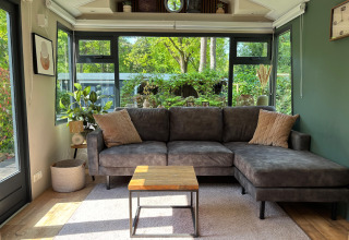 Gemütliches Wohnzimmer im Veluwe Chalet mit grau L-förmigem Sofa, Pflanzen und Ausblick auf den Garten.