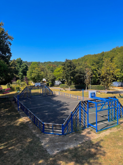 Cancha deportiva con valla azul en Camping Le Périgord en Nouvelle-Aquitaine, Francia, rodeada de naturaleza.