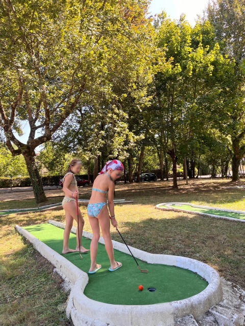 Dos niños en traje de baño juegan al minigolf bajo árboles soleados en un parque vacacional en Francia.
