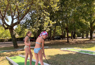 Dos niños en traje de baño juegan al minigolf bajo árboles soleados en un parque vacacional en Francia.