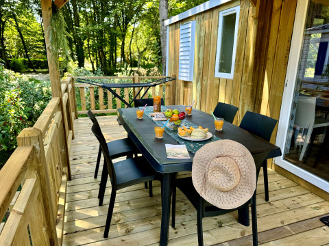 Terrazza in legno con tavolo apparecchiato per sei, frutta, succhi e cappello al Camping Le Périgord, Nouvelle-Aquitaine.