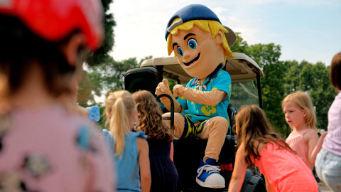 Children gather around a cheerful mascot in a cap and Hawaiian shirt at a glamping site on a sunny day.