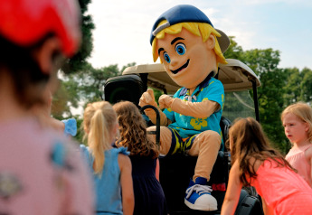 Children gather around a cheerful mascot in a cap and Hawaiian shirt at a glamping site on a sunny day.