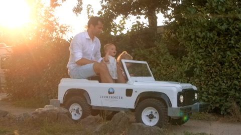 A man and child ride a small Leukermeer car at a glamping site, with a sunset shining in the background.