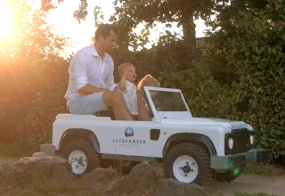 Un homme et un enfant roulent dans une petite voiture Leukermeer au glamping, soleil couchant en arrière-plan.