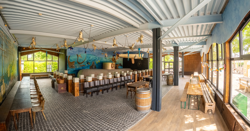 Luminoso salón común glamping con decoración marina, bancas de madera y amplias ventanas panorámicas.