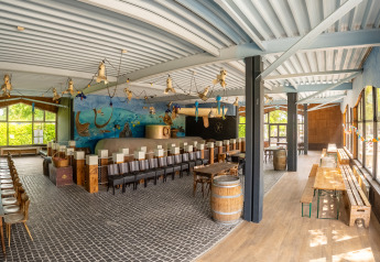 Grande salle commune de glamping lumineuse, décor marin, grandes fenêtres et longues tables en bois.