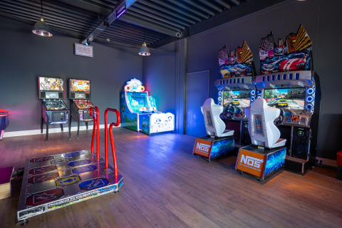 Sala de juegos arcade luminosa en un alojamiento glamping con simuladores de coches y máquinas recreativas.