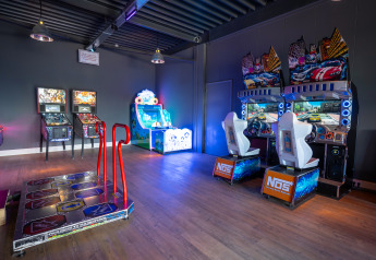 Sala de juegos arcade luminosa en un alojamiento glamping con simuladores de coches y máquinas recreativas.