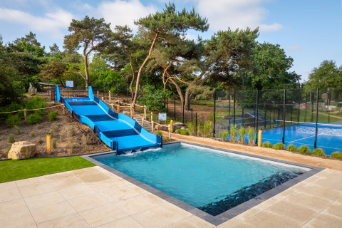 Piscina al aire libre con tobogán azul y pista deportiva al lado de alojamiento glamping rodeado de árboles.