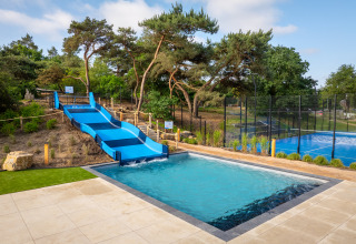 Buitenzwembad met blauwe glijbaan en sportveld bij een glampingverblijf in een bosrijke omgeving.