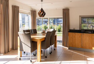 Comedor con grandes ventanales, cortinas beige y muebles de madera en Wellness Lodge XL, Hof van Salland.