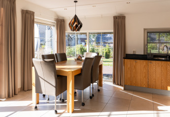 Comedor con grandes ventanales, cortinas beige y muebles de madera en Wellness Lodge XL, Hof van Salland.