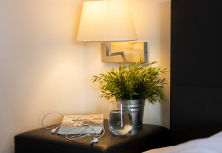 Mesa de noche acogedora con lámpara, vaso de agua, libro, planta y auriculares en Wellness Lodge XL.