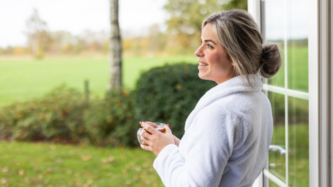 Frau im weißen Bademantel genießt Tee am Fenster der Wellness Lodge XL in Hof van Salland, Niederlande.