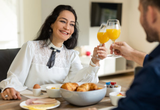 Couple enjoys breakfast with orange juice in Wellness Lodge XL at Hof van Salland, Netherlands, smiling.