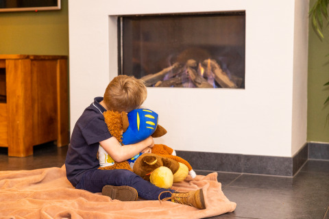 Un niño sentado sobre una manta frente a la chimenea en Wellness Lodge XL, Hof van Salland, Países Bajos, abraza peluches.