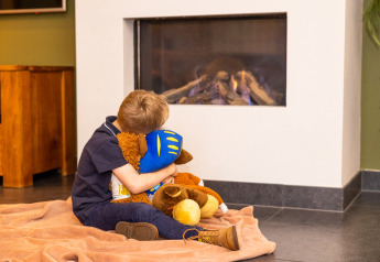 Un niño sentado sobre una manta frente a la chimenea en Wellness Lodge XL, Hof van Salland, Países Bajos, abraza peluches.