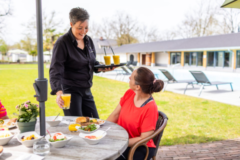 Donna fa colazione all’aperto mentre una cameriera serve succo fresco nella Wellness Lodge XL a Hof van Salland.
