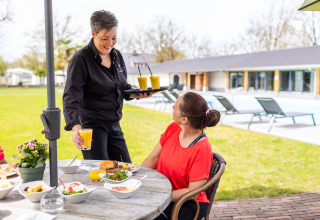 Una mujer desayuna al aire libre mientras una camarera sirve jugo fresco en Wellness Lodge XL, Hof van Salland, Países Bajos.