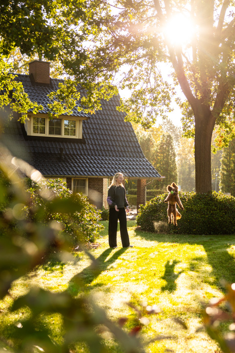 Mujer juega con un perro saltando frente a una casita en el sol en Wellness Lodge XL, Hof van Salland.