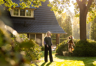 Mujer juega con un perro saltando frente a una casita en el sol en Wellness Lodge XL, Hof van Salland.