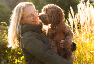 Mujer rubia con gafas sostiene un perro marrón frente a Wellness Lodge XL en Hof van Salland, Países Bajos.
