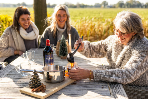 Three women share mulled wine outdoors at Wellness Lodge XL, Hof van Salland, Netherlands, in sunlight.