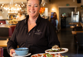 Mujer con camisa negra sirviendo comida en Wellness Lodge XL de Hof van Salland, ambiente acogedor de restaurante.