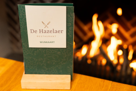 Carta del restaurante De Hazelaer sobre una mesa frente a una chimenea en Wellness Lodge XL, Hof van Salland.