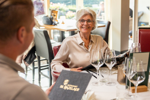 Anziana donna sorride al tavolo di un ristorante con menu in mano, Wellness Lodge XL, Hof van Salland, Paesi Bassi.