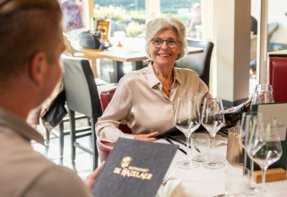 Mujer mayor sonríe en una mesa de restaurante con un menú en mano en Wellness Lodge XL, Hof van Salland, Países Bajos.