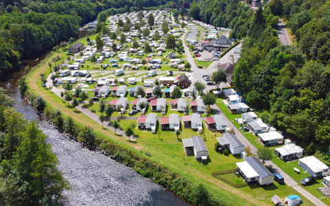 Luftaufnahme eines Glampingplatzes mit Wohnwagen und Hütten am Fluss, umgeben von grüner Natur.