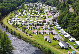 Luftaufnahme eines Glampingplatzes mit Wohnwagen und Hütten am Fluss, umgeben von grüner Natur.
