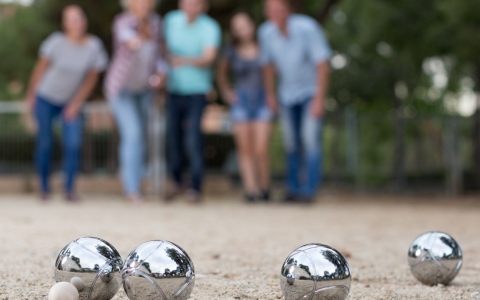 Vier Personen spielen Boule beim Glamping, Fokus auf glänzenden Kugeln auf sandigem Boden im Freien.
