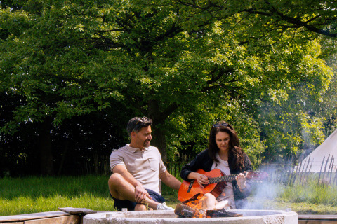 Et par nyder et bål, mens kvinden spiller guitar, omgivet af grønne træer på glampingpladsen.