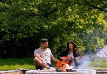 Ein Paar entspannt am Lagerfeuer, die Frau spielt Gitarre, beim Glamping inmitten grüner Natur.