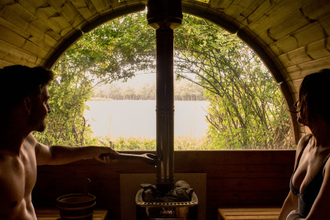 Pareja relajándose en una sauna acogedora con vista al lago desde un alojamiento de glamping en la naturaleza.